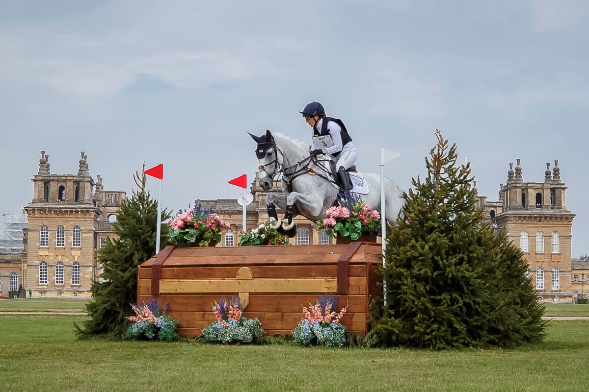 A women jumping on a horse over a jump with Blenheim Palace in the background.
