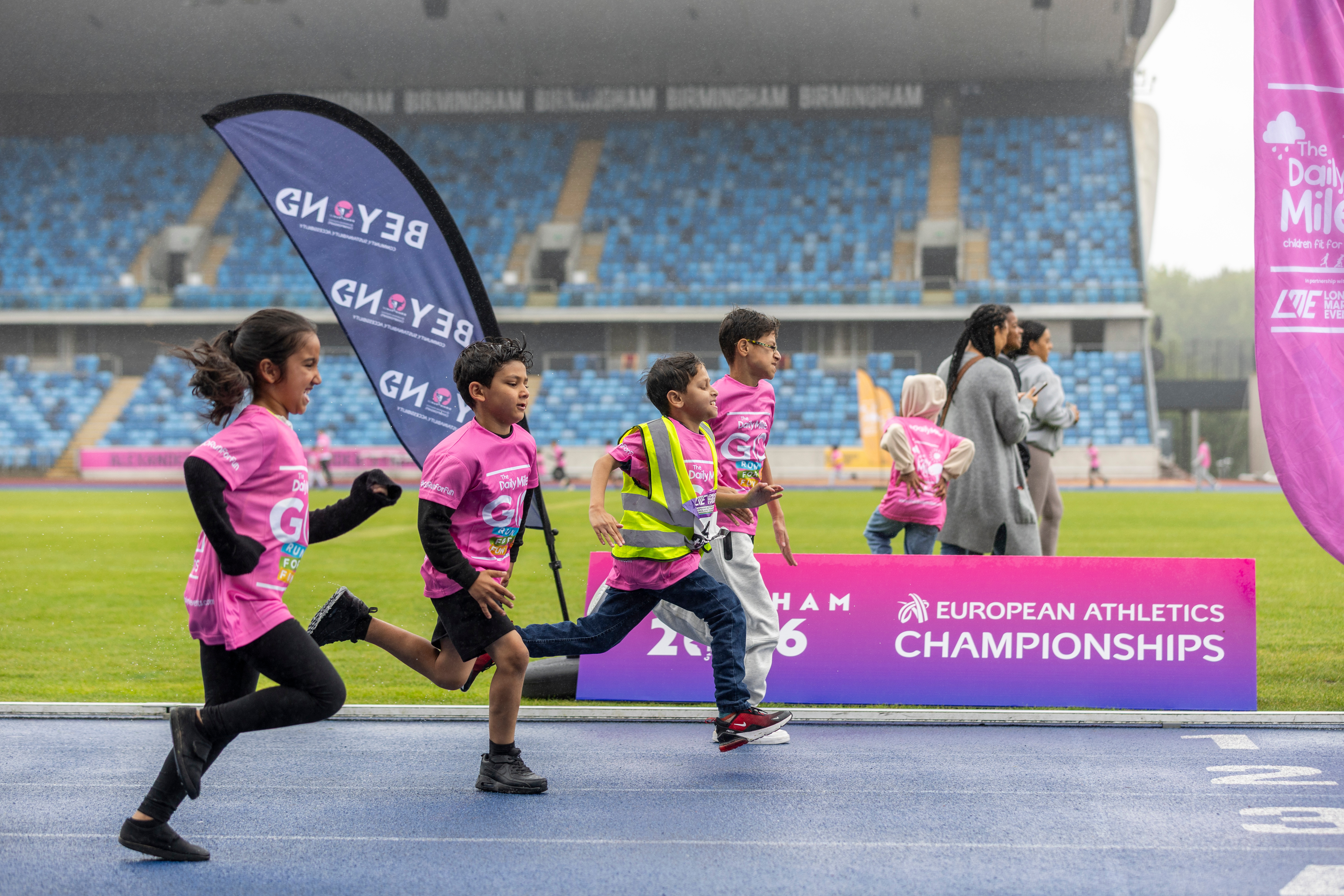 Children run on a running track towards the finish line in the pouring rain. The side on shot shows the Birmingham Alexander Stadium behind them.