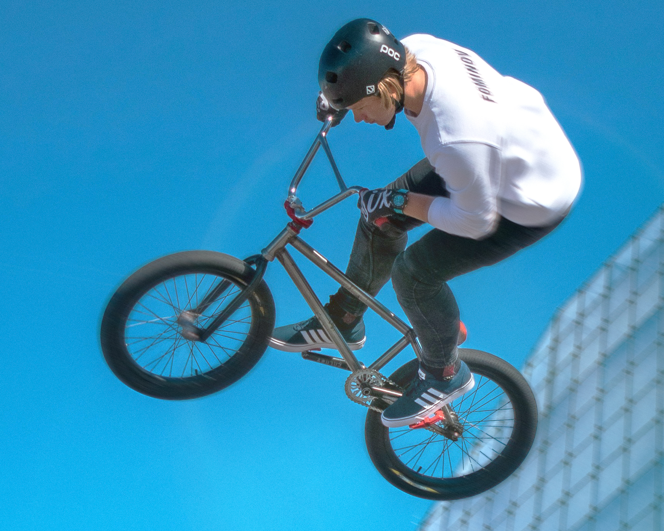 A BMX rider wearing a black helmet, white long-sleeved top and dark trousers performs an aerial trick on a BMX bike against a bright blue sky.