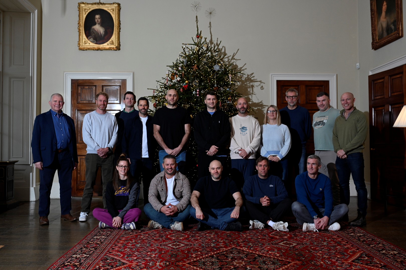 Group photo of people standing and sitting in front of a decorated Christmas tree in a formal room.
