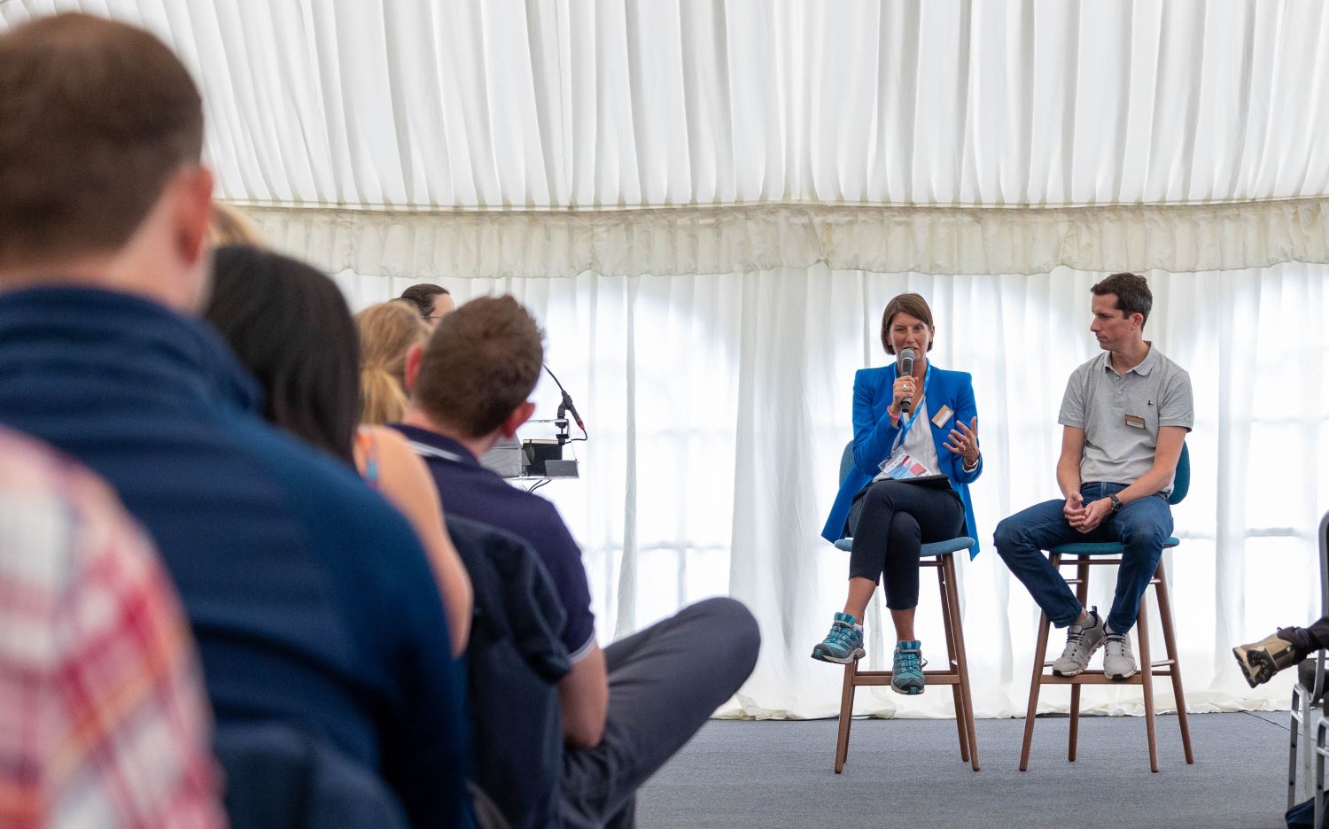 Esther Britten sits on a high stool at the front of an audience speaking to them whilst holding a microphone.