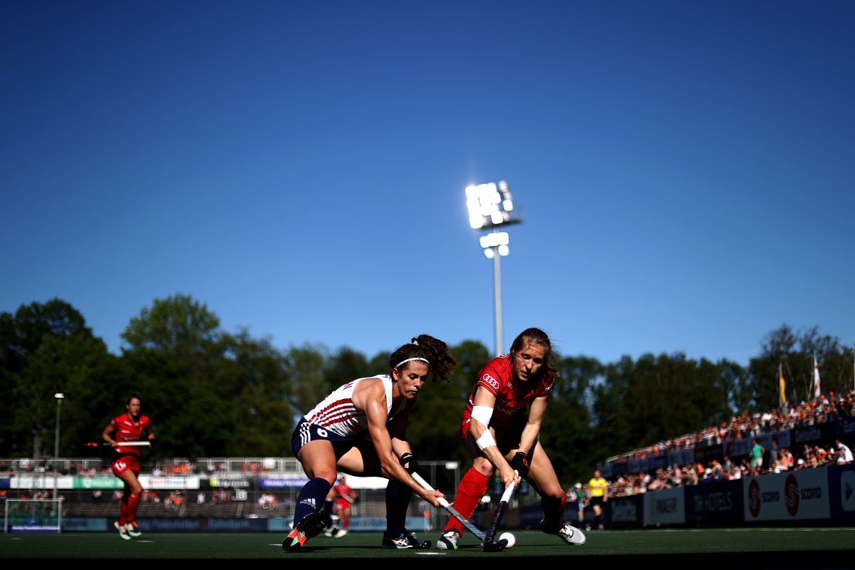Two Hockey players compete for the ball in front of a crowd. The sky is blue and a flood light is in view above the stands.