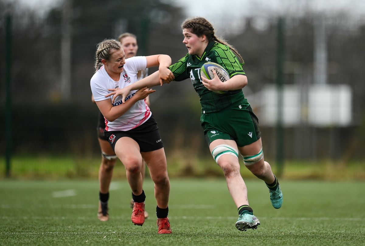 Irish player Jemima fends off an opposing player in a match whilst running with the rugby ball under her left arm.