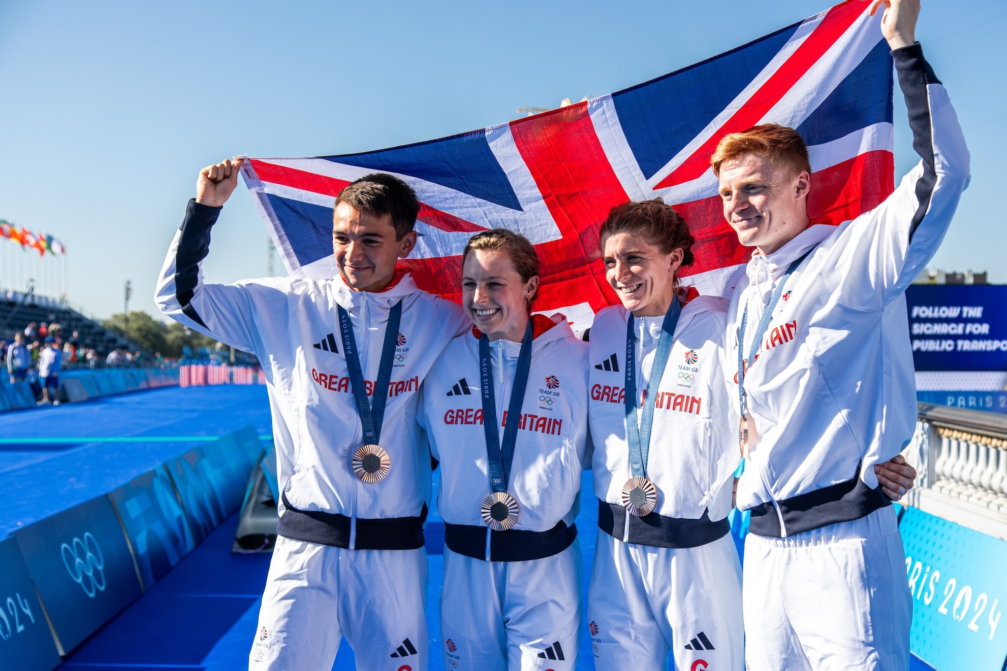 Team GB Team Triathlon athletes celebrating their medal at the Paris 2024 Olympic Games, holding the Union Jack flag 