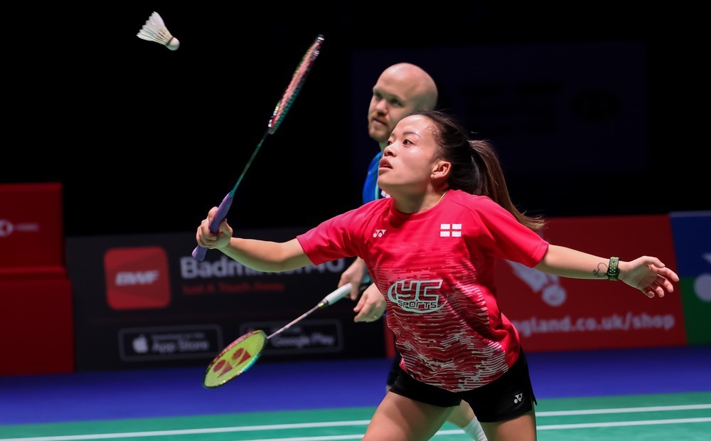 A female athlete, wearing a red t-shirt playing para-badminton. In the background is her doubles partner, a male athlete wearing a blue t-shirt