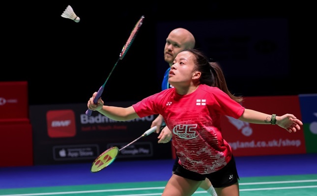 A female athlete, wearing a red t-shirt playing para-badminton. In the background is her doubles partner, a male athlete wearing a blue t-shirt