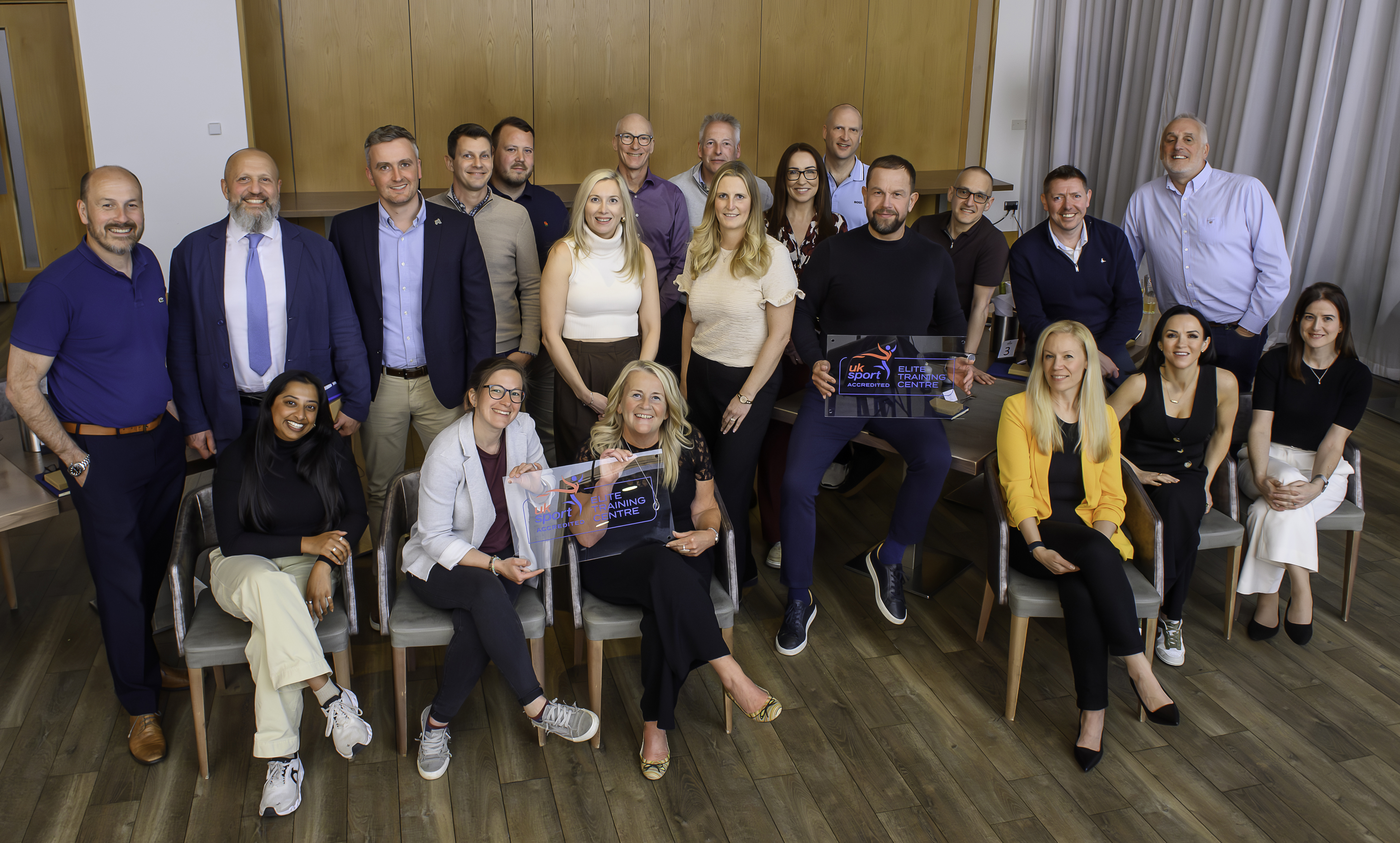 A group of people pose for a formal photo indoors. Some are seated and others standing, with two people holding glass awards, in front of a wood‑panelled wall.