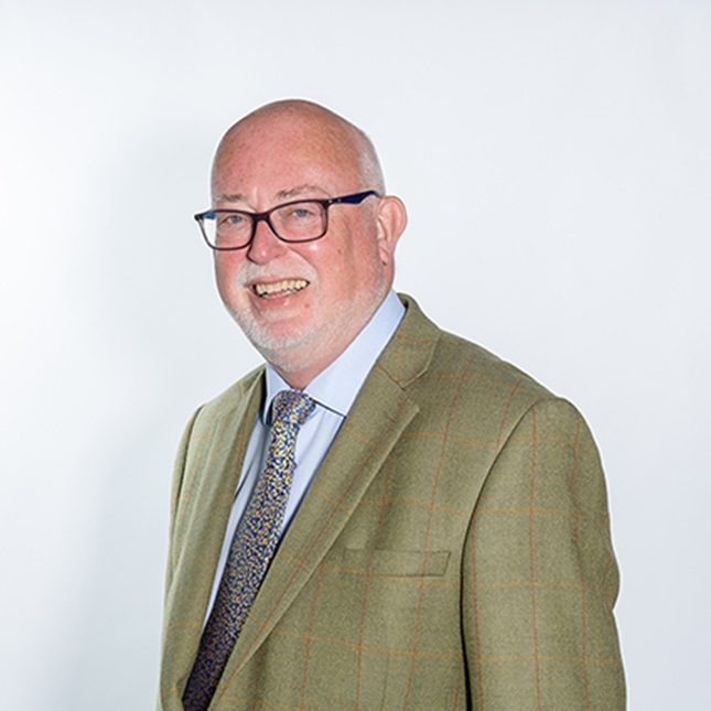 A man in a shirt and tie and green jacket - wearing glasses he has a smile with a white beard.