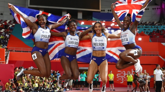 A team of British relay athletes celebrating after a race. Jumping in the air and holding a Union Jack flag