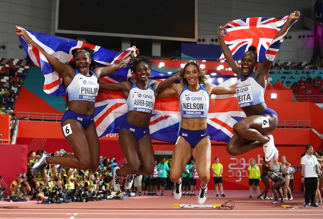 A team of British relay athletes celebrating after a race. Jumping in the air and holding a Union Jack flag