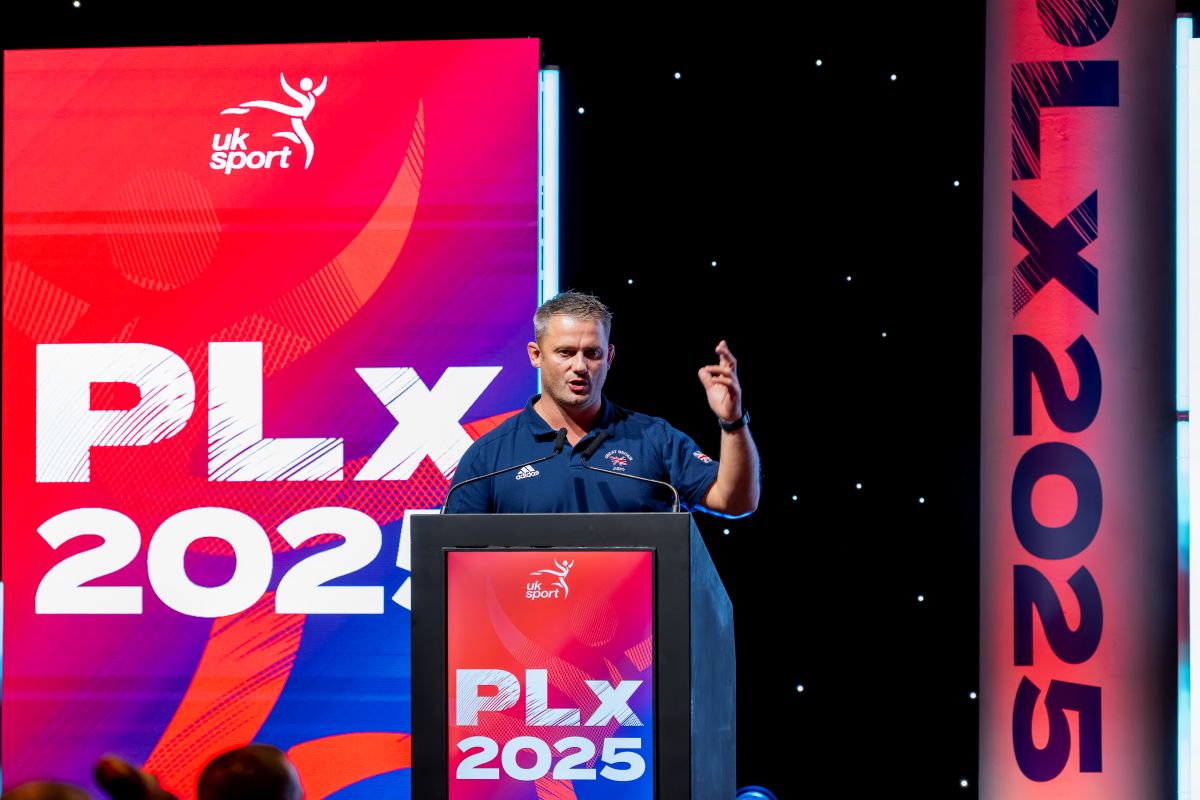 Speaker at a podium during UK Sport PLx 2025 event with bold branding on stage backdrop.