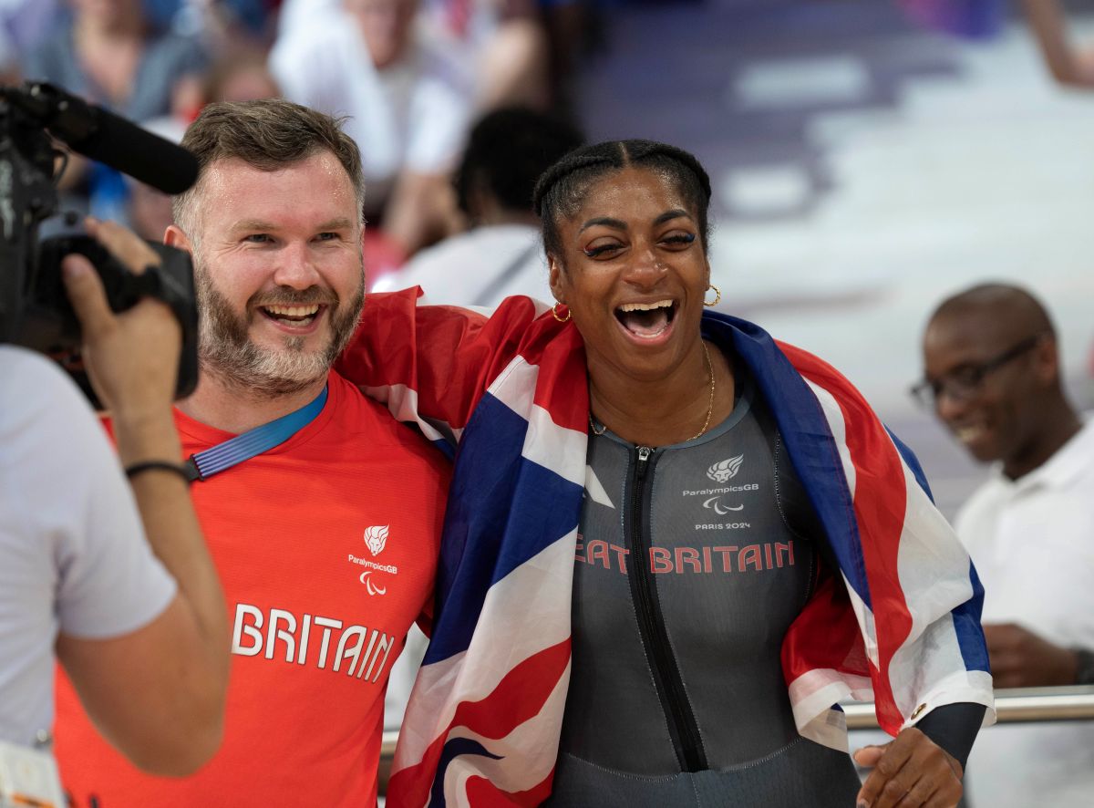 Kadeena Cox, in a Great Britain navy cycling skinsuit is held up by her coach in a red t shirt. They celebrate with a Union Jack flag at the Paris 2024 Paralympic Games.