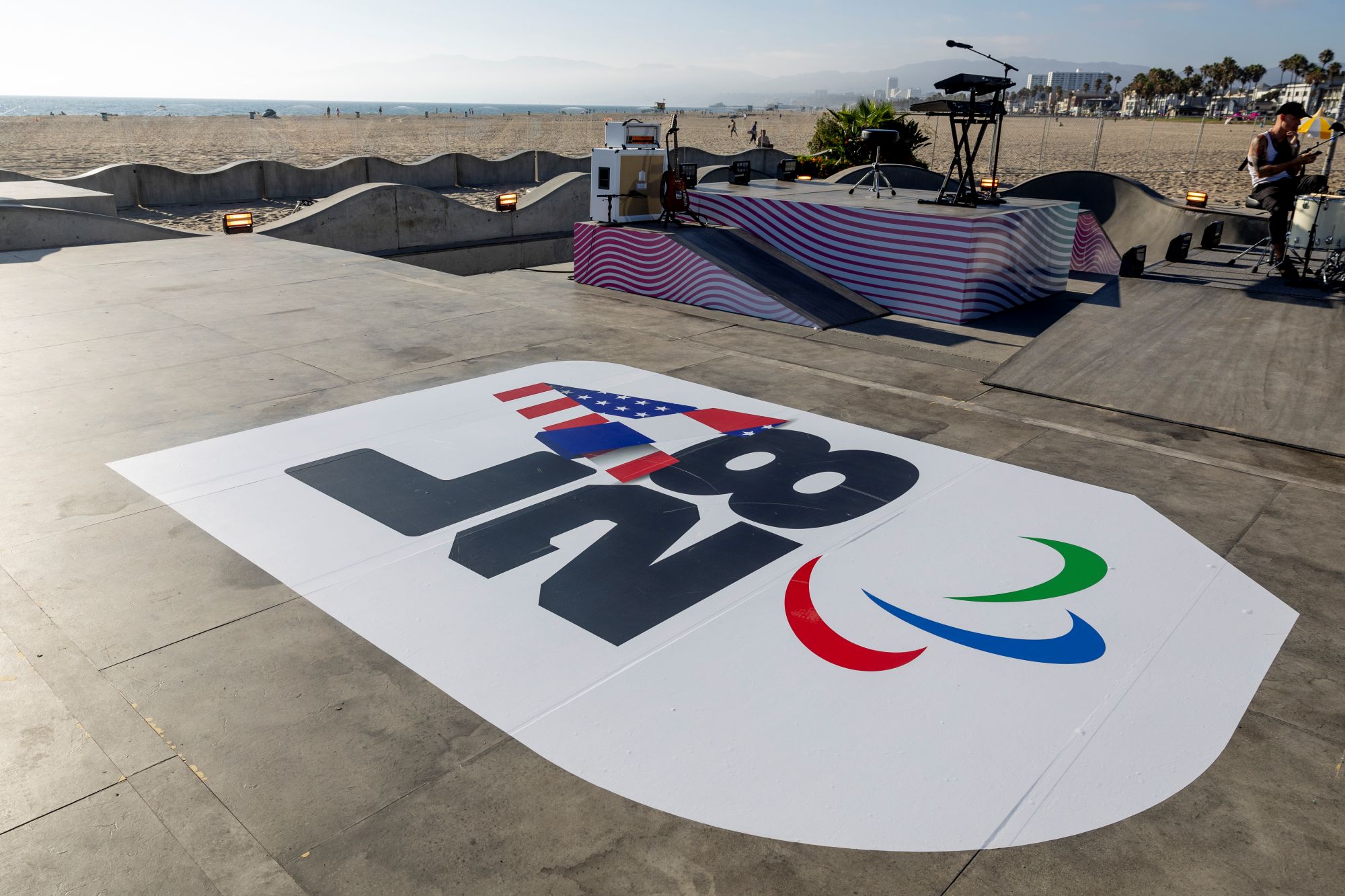 The LA28 logo is emblazed on the floor in a logo black text on white background. It features the USA flag and Paralympics Agitos logo of a red, green and blue flash of colour. It is in a skatepark on a beach looking out towards the sea.