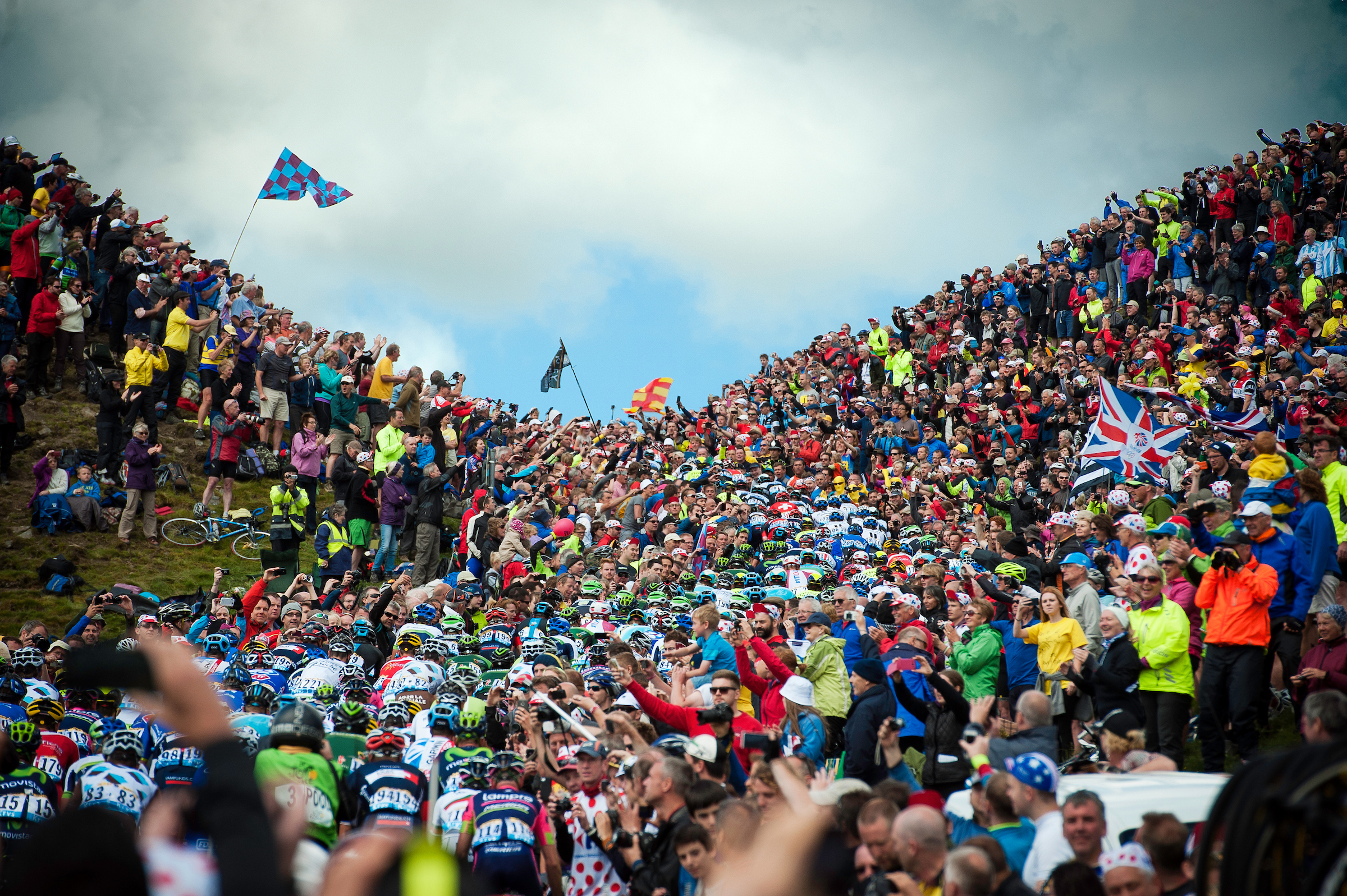 A large crowd lines both sides of a steep hillside, cheering and taking photos as a dense group of cyclists in colorful jerseys and helmets rides through the narrow gap. Bright flags, including a Union Jack and patterned banners, are visible among the spectators. The sky is partly cloudy, adding contrast to the vibrant scene of the cycling event.