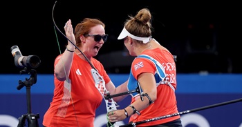 Two people embrace each other smiling during a sporting competition. One holds an bow whilst embracing - celebrating a good shot.