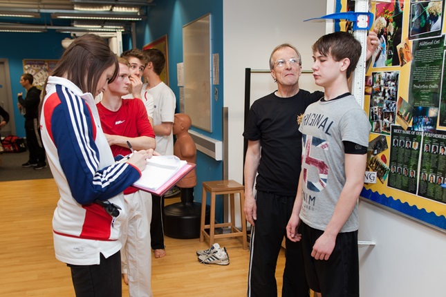 A child having their height measured by an adult whilst an observer makes notes on a clipboard