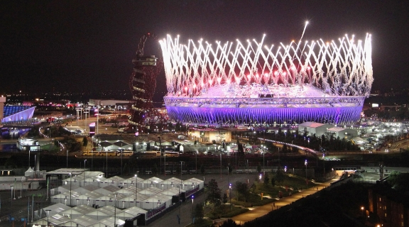 View of a city skyline at night with a sports stadium in the background lit by fireworks