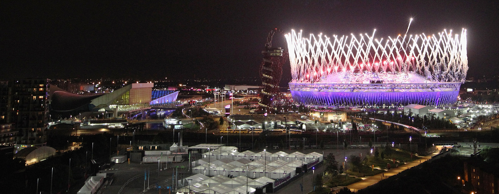 View of a city skyline at night with a sports stadium in the background lit by fireworks