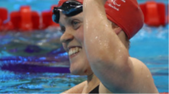 Team GB athlete Ellie Simmonds in a swimming pool during competition, wearing a red swimming cap, black swimsuit and goggles and smiling