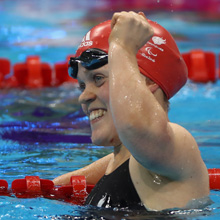 Team GB athlete Ellie Simmonds in a swimming pool during competition, wearing a red swimming cap, black swimsuit and goggles and smiling