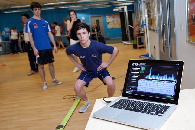 A close up of a laptop computer screen showing data and charts. Young people stood in the background performing exercises