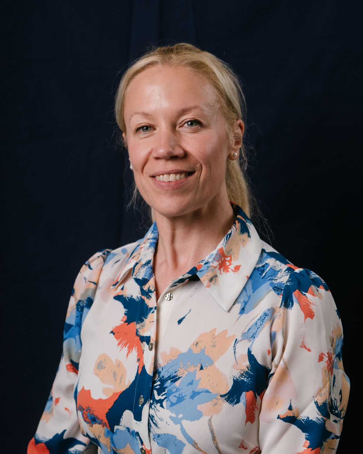 A portrait picture of Kate Baker, Director of Performance and People at UK Sport, she smiles at the camera in a colourful floral shirt of blues, reds and cream.