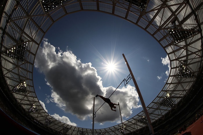 An athlete leaps over the bar at the top of their pole vault attempt in the London Stadium, the image looking up at the sun shining and blue sky above.