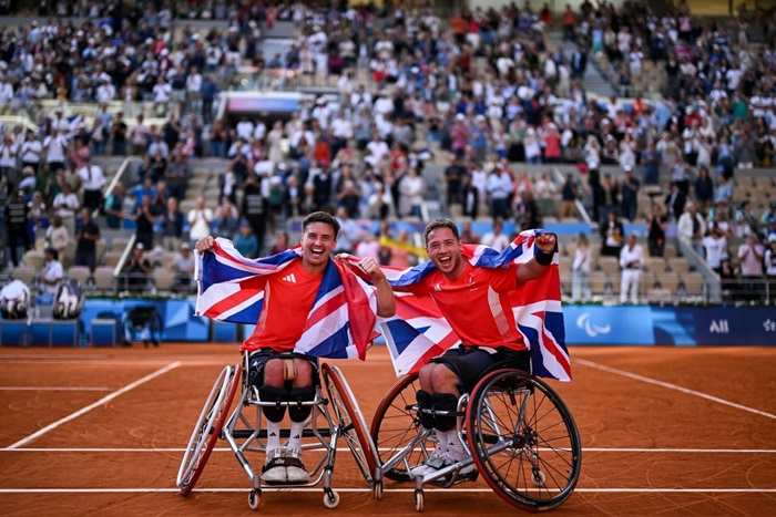 Alfie Hewett and Gordon Reid celebrate in Roland Garros, raising the Union Jack flag over their heads as they pose for the camera. The crowd can be seen celebrating behind them.
