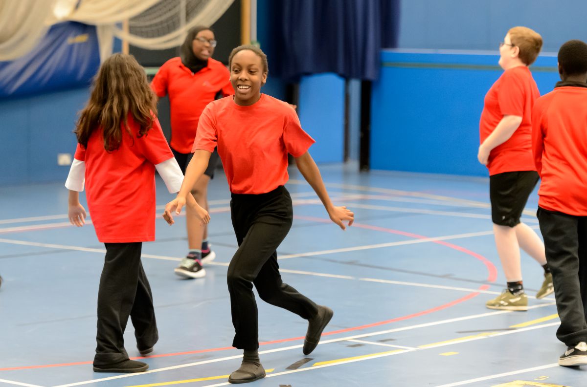 Children play together in a sports hall, smiling as the skip around.