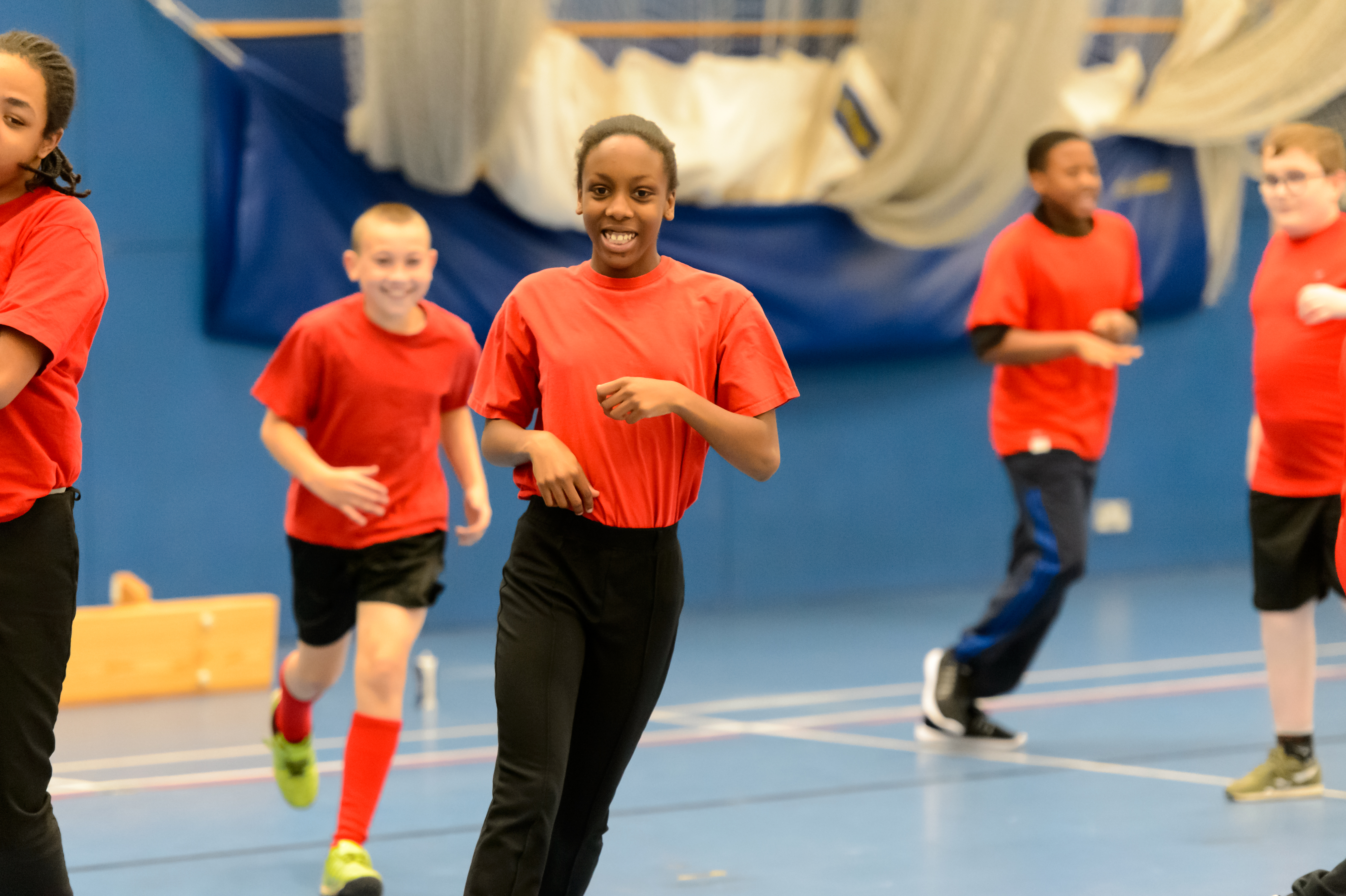 Children run around an indoor sports hall, smiling