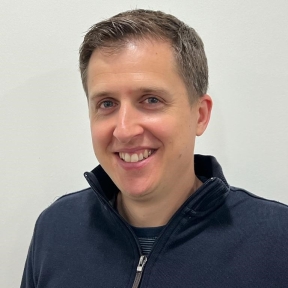 Portrait of man with short light brown hair, smiling, wearing a navy zip-up top