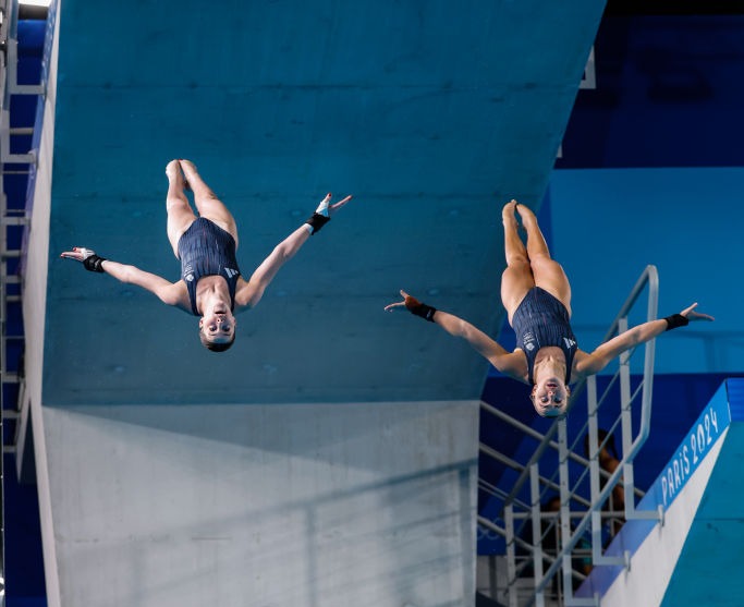 Two women dive off the top of a diving board into a pool.