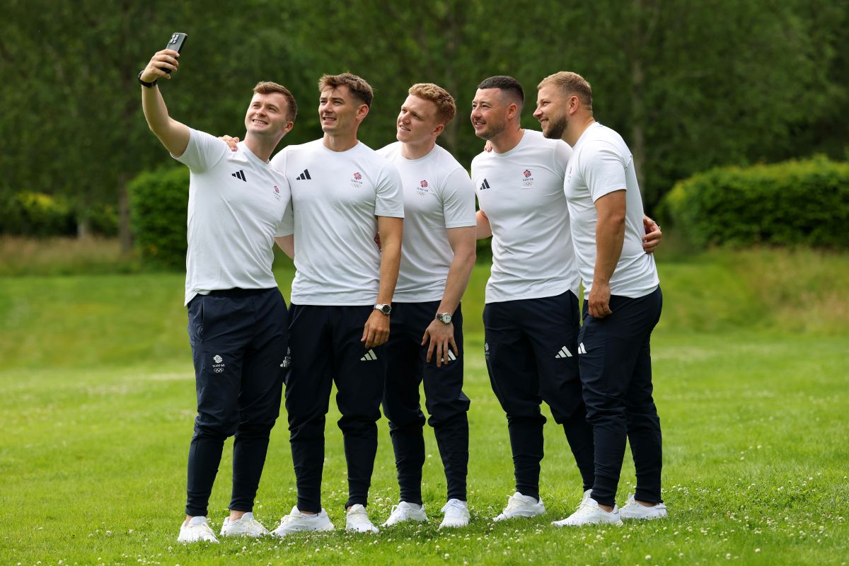 Five men in white t shirts and blue trousers take a selfie in a park.