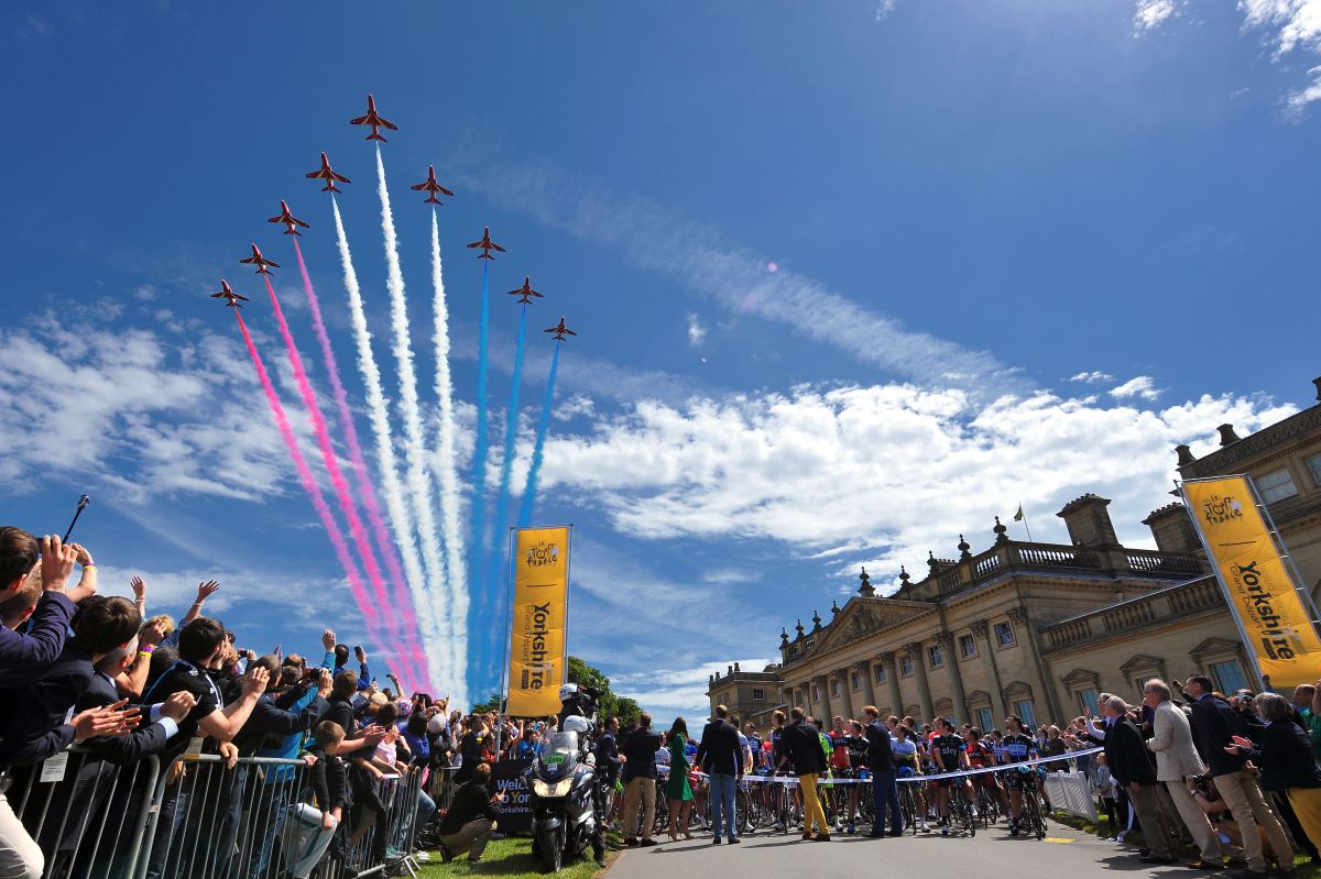 Crowds line the street in front of a grand classical building in Yorkshire, watching seven aircraft fly in formation with red, white, and blue smoke trails during the Tour de France 2014 Grand Départ. Cyclists and officials gather near the starting line beneath tall yellow 'Yorkshire' banners.
