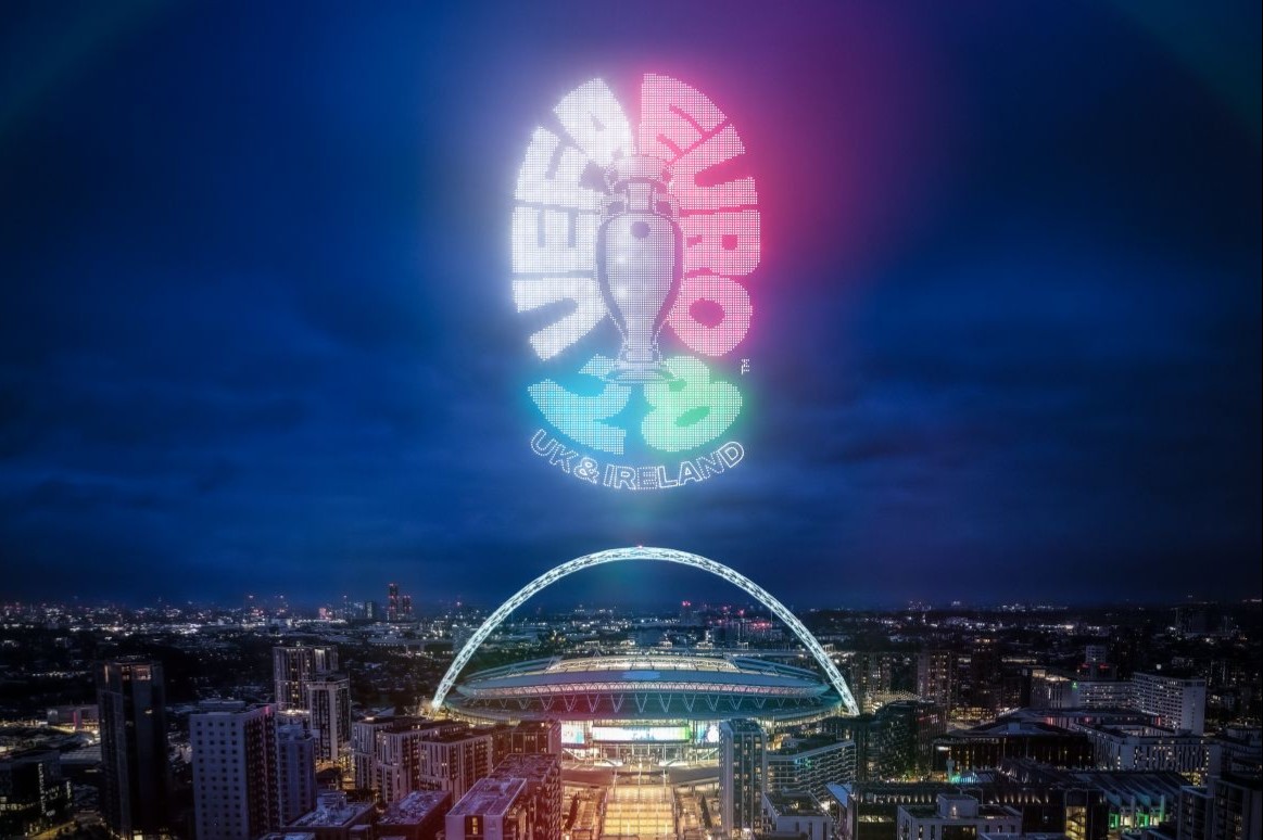 Night-time aerial view of Wembley Stadium in London, illuminated with bright lights, with the UEFA EURO 2028 logo projected in vibrant colours above the stadium. The logo includes the tournament trophy and text reading ‘UEFA EURO 2028 UK & Ireland’ against a dark blue sky.”