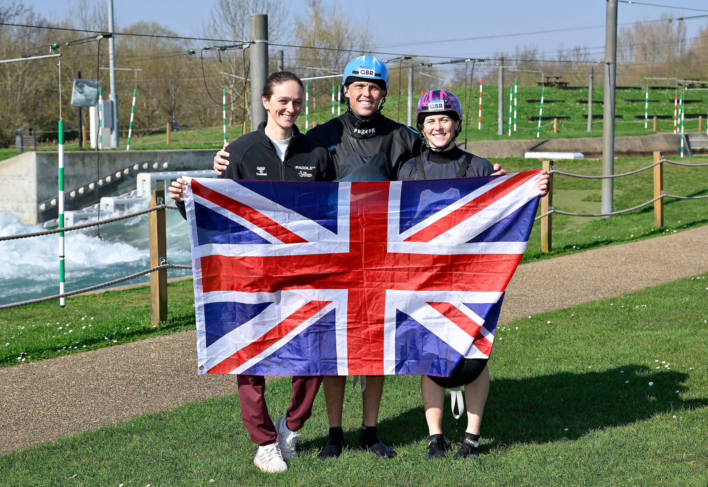 Three slalom paddlers hold a Union flag beside a white water course.